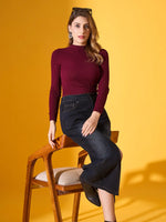 A woman wears the 98degreenorth Rosewood Ribbed High-Neck Sweater, sitting on a wooden chair against yellow.