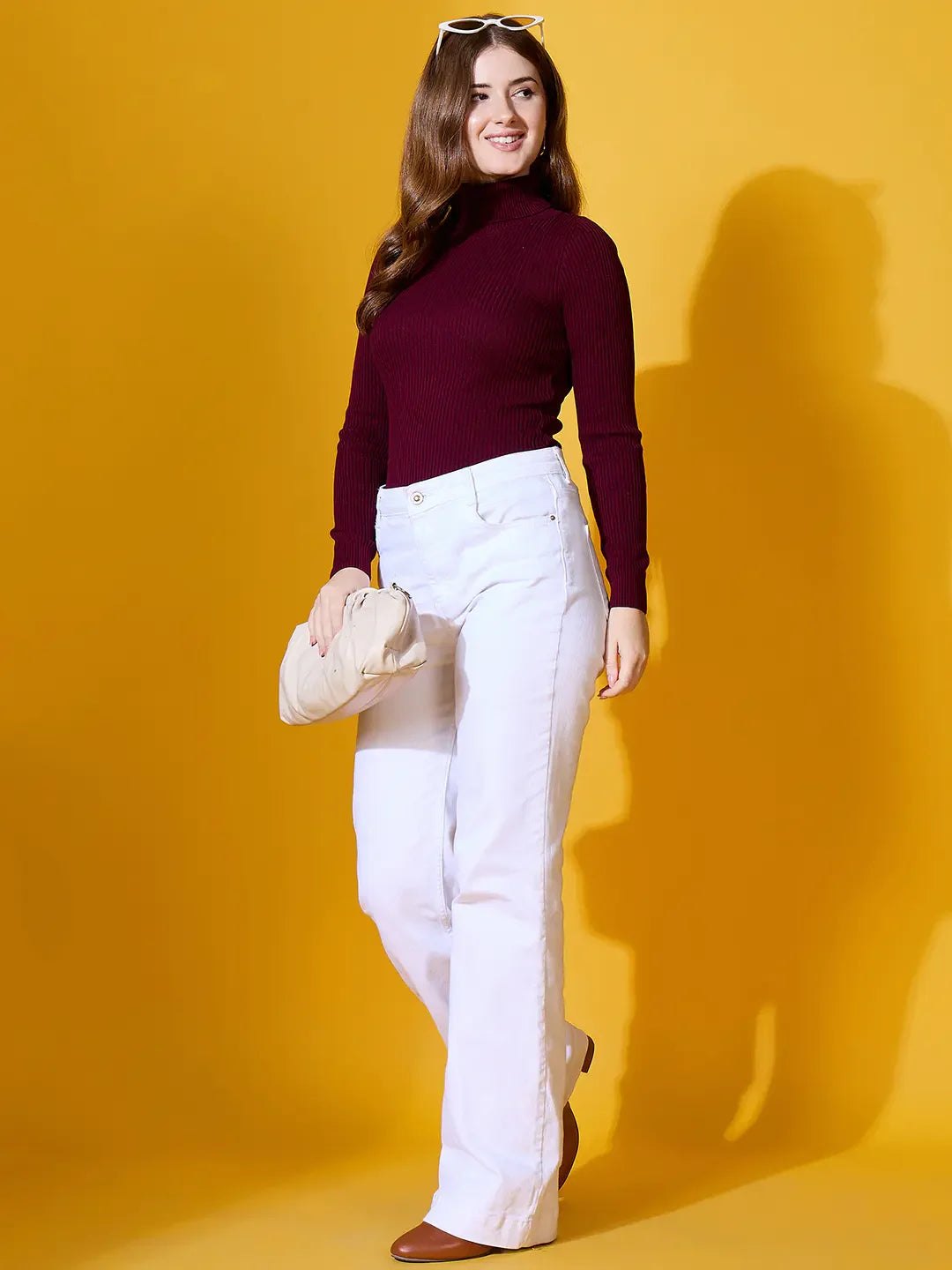A woman in a 98degreenorth Rosewood Ribbed Turtle-Neck Pullover poses with a cream clutch against a yellow background.
