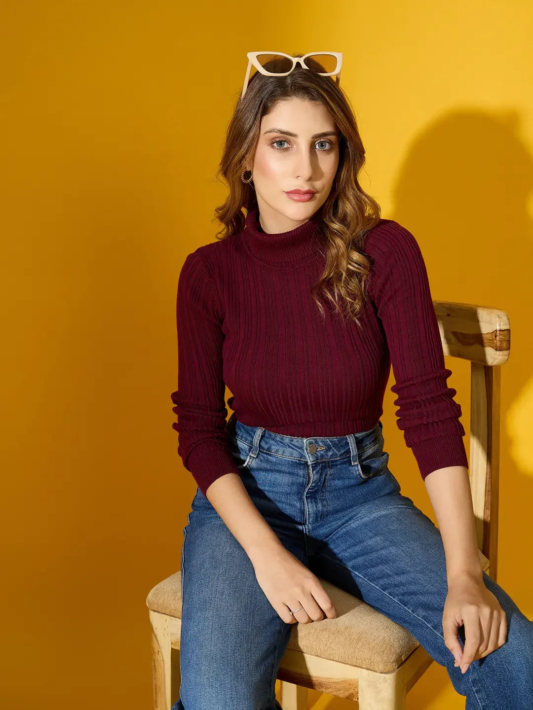 Woman in a 98degreenorth rosewood ribbed turtleneck sweater and blue jeans sits on a chair, yellow background.