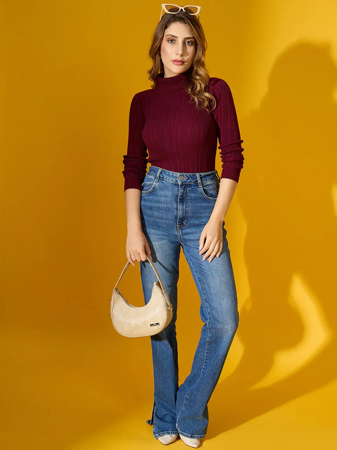Woman in 98degreenorth Rosewood Ribbed Turtleneck Sweater and jeans poses with a beige handbag against a yellow background.