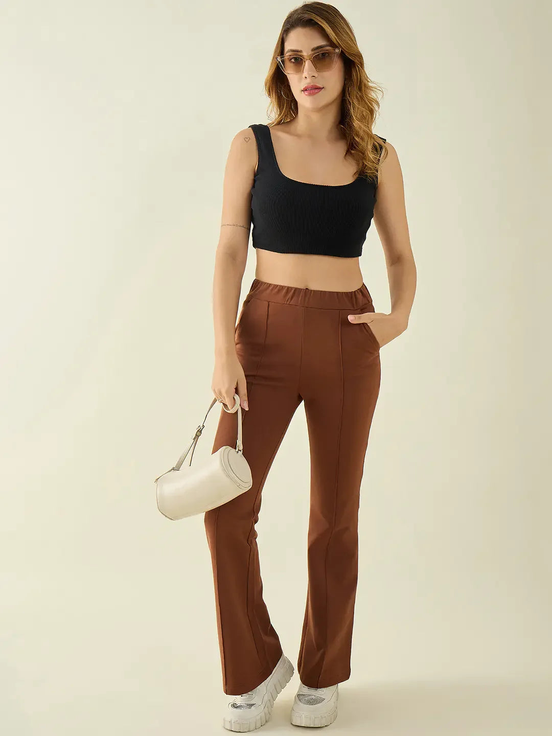 Woman in sunglasses wears a black crop top, 98degreenorth rust flared bootcut trousers, white sneakers, and a white handbag.