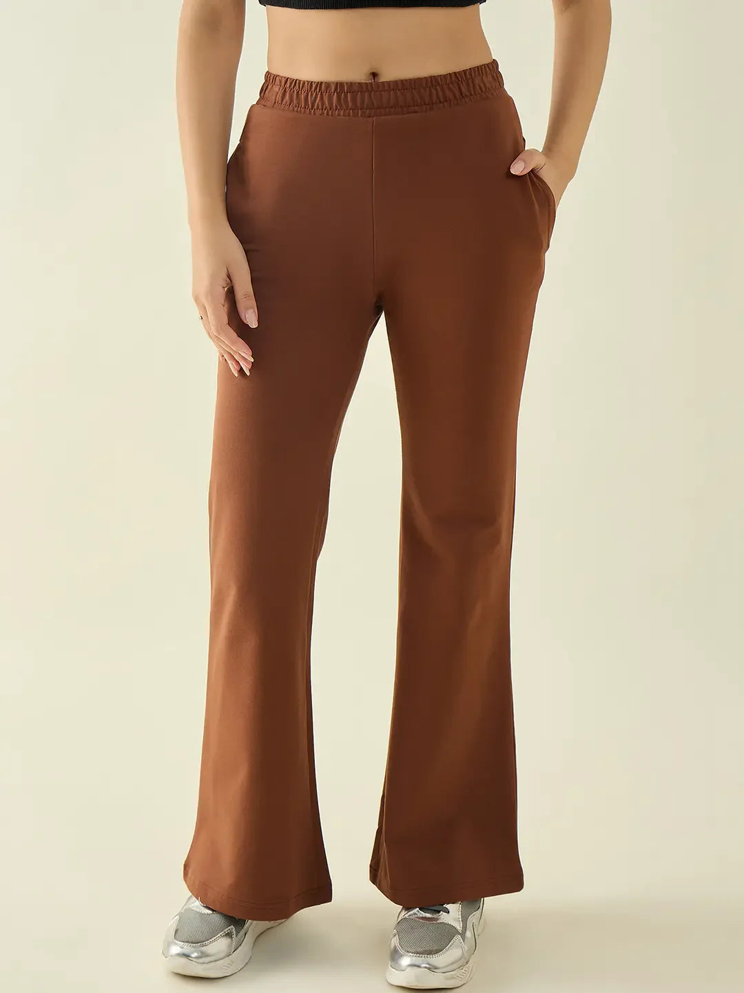 Woman in 98degreenorth Rust Flared Bootcut Trousers, black crop top, and silver sneakers stands hands in pockets.