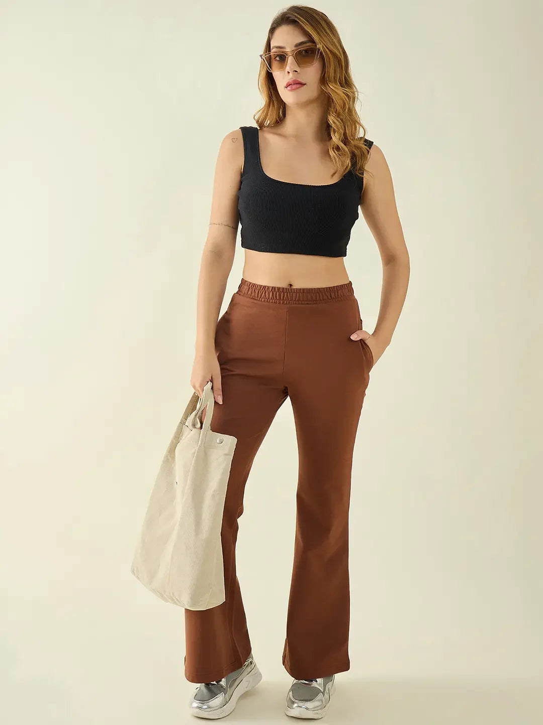 Woman in a black crop top, 98degreenorth rust bootcut trousers, sunglasses, sneakers & beige tote bag.
