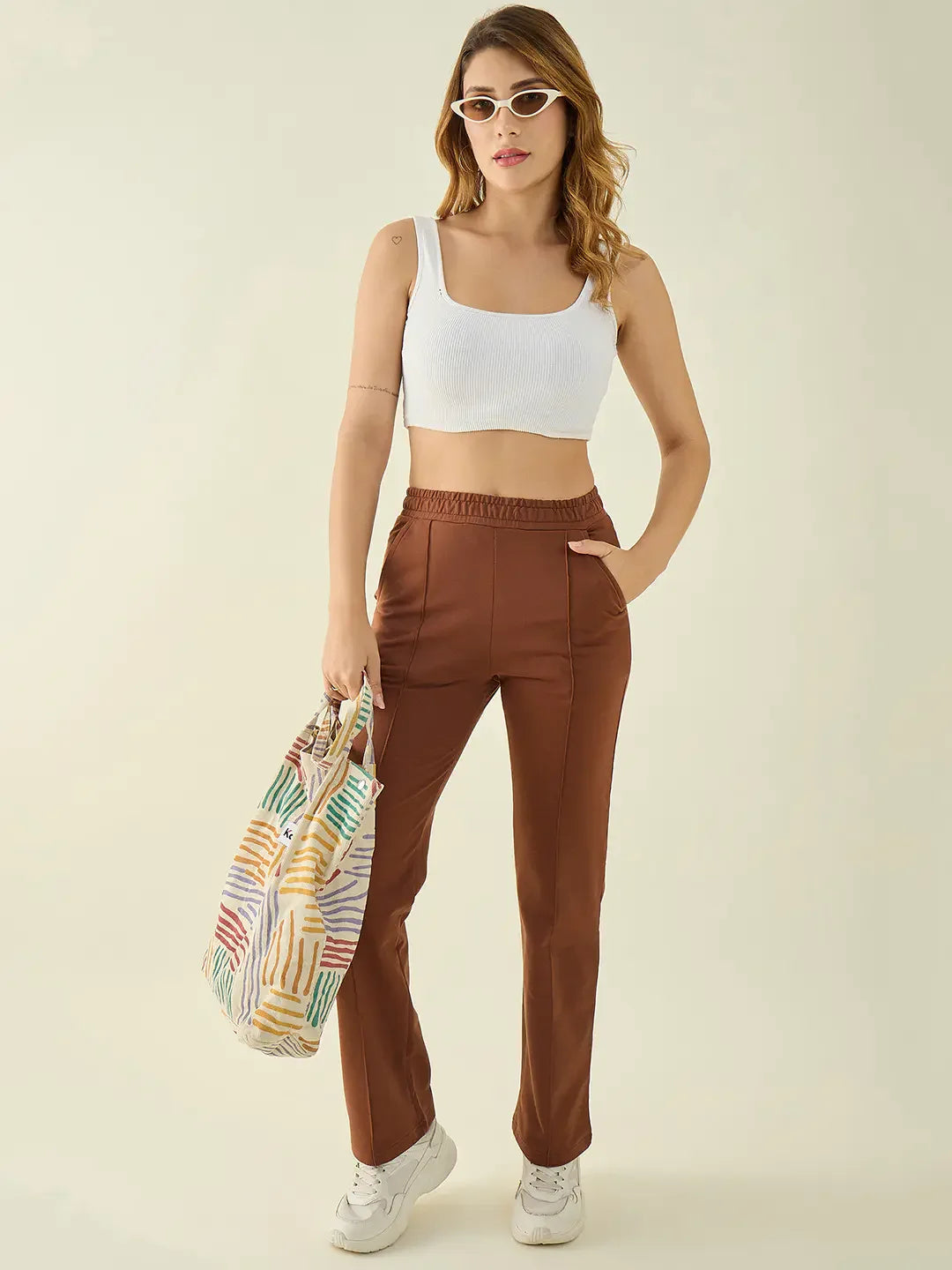 Woman in a white crop top, 98degreenorth rust straight fit trousers, white sneakers, sunglasses, and a striped tote.
