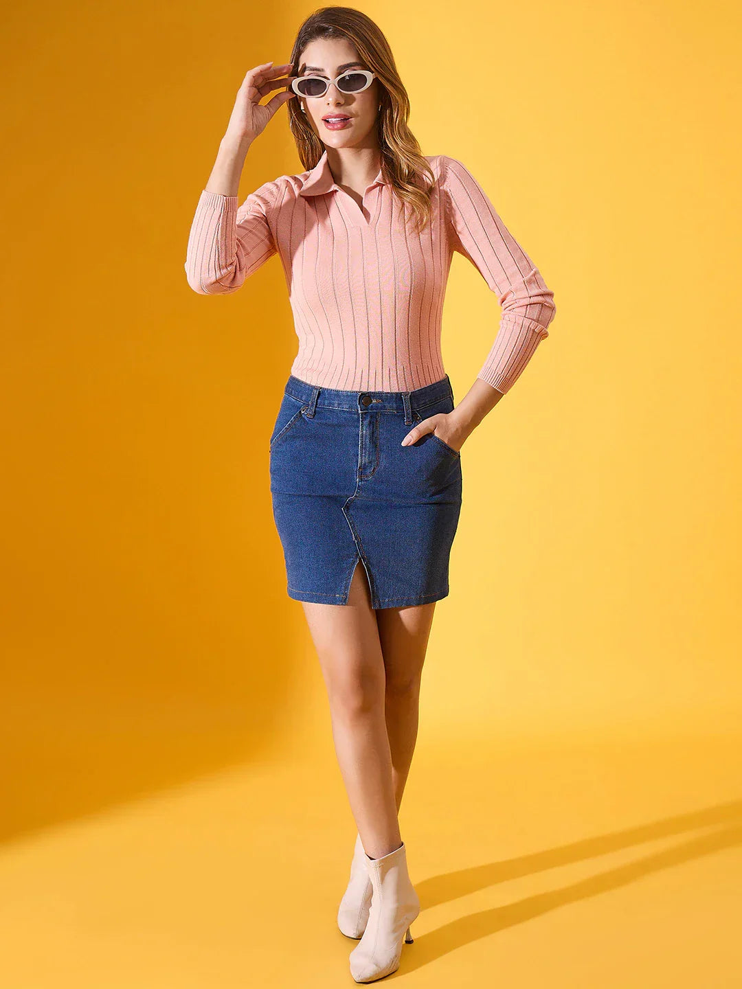 Woman in sunglasses wears a 98degreenorth salmon ribbed collar neck sweater, denim skirt, and white boots.