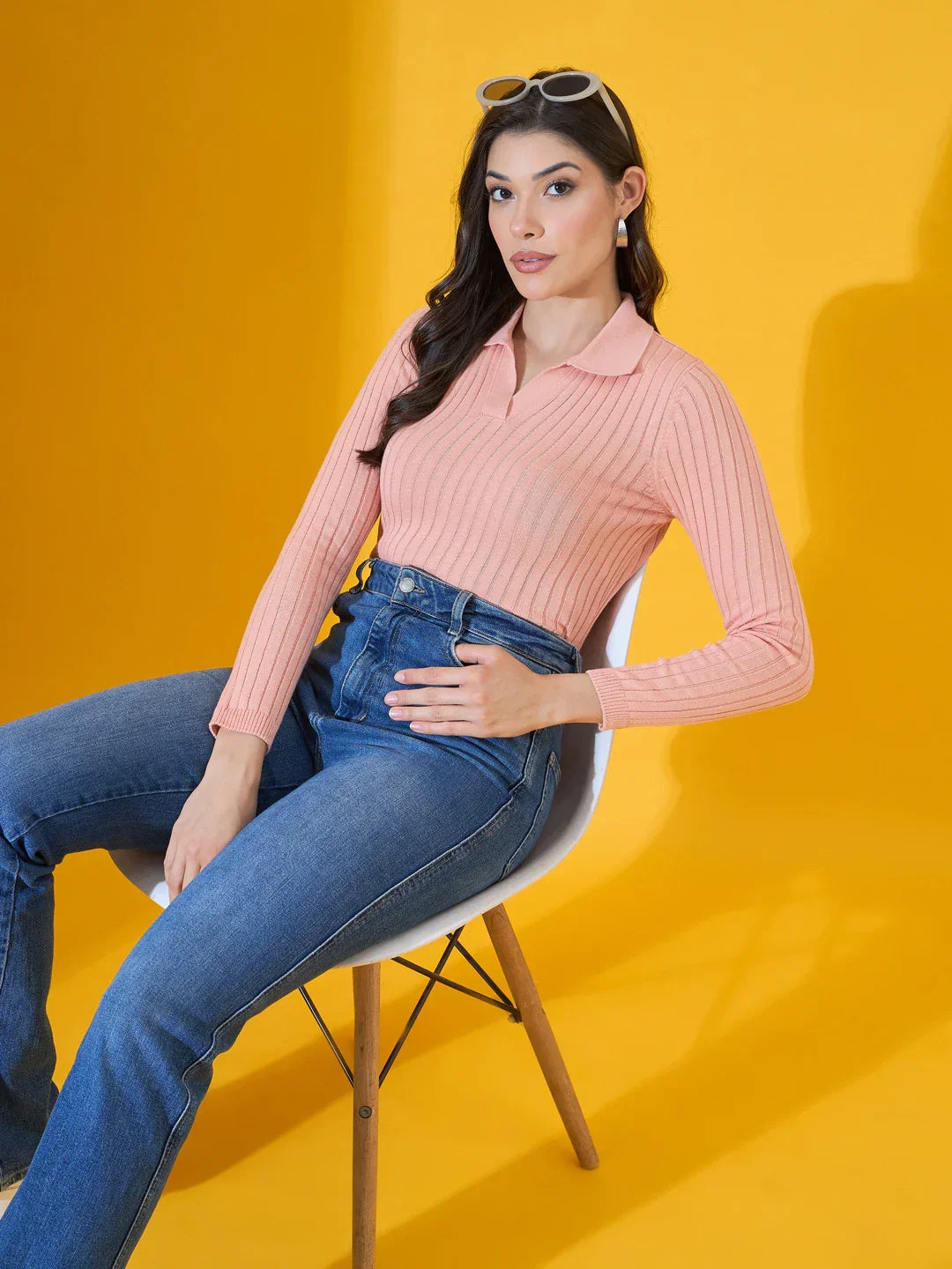 A woman in a 98degreenorth salmon ribbed slim fit sweater sits on a white chair against a yellow backdrop.