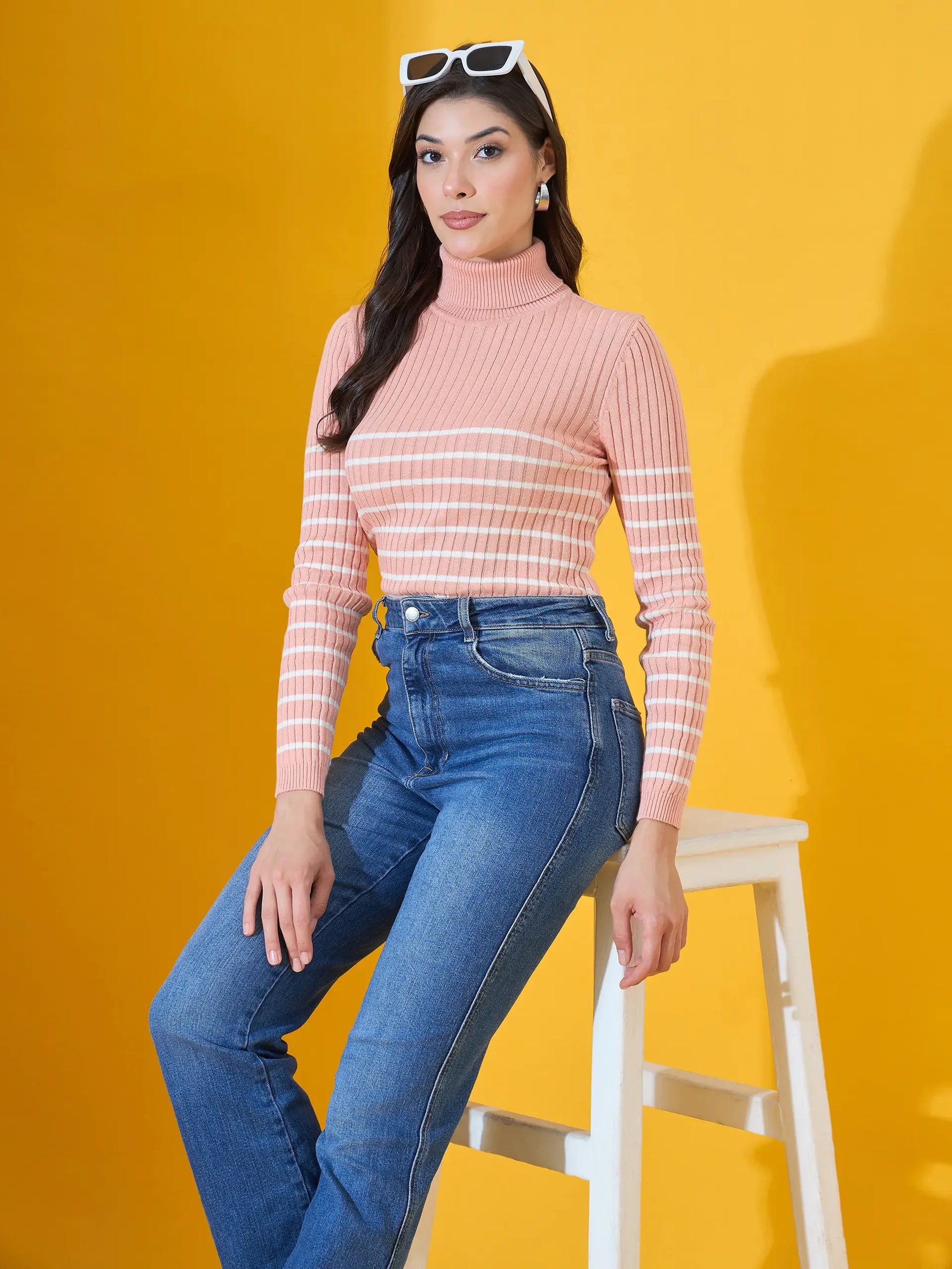 Woman wears 98degreenorth Women’s Salmon Ribbed Turtleneck Sweater, sitting on a white stool with yellow background.