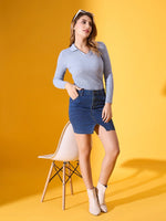Woman in a 98degreenorth sky blue ribbed collar sweater and denim skirt poses by a white chair with boots and sunglasses.