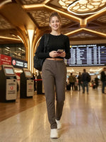 A woman in 98degreenorth's Olive Straight Fit Cotton Trackpants walks at the airport, phone in hand, flight boards behind.