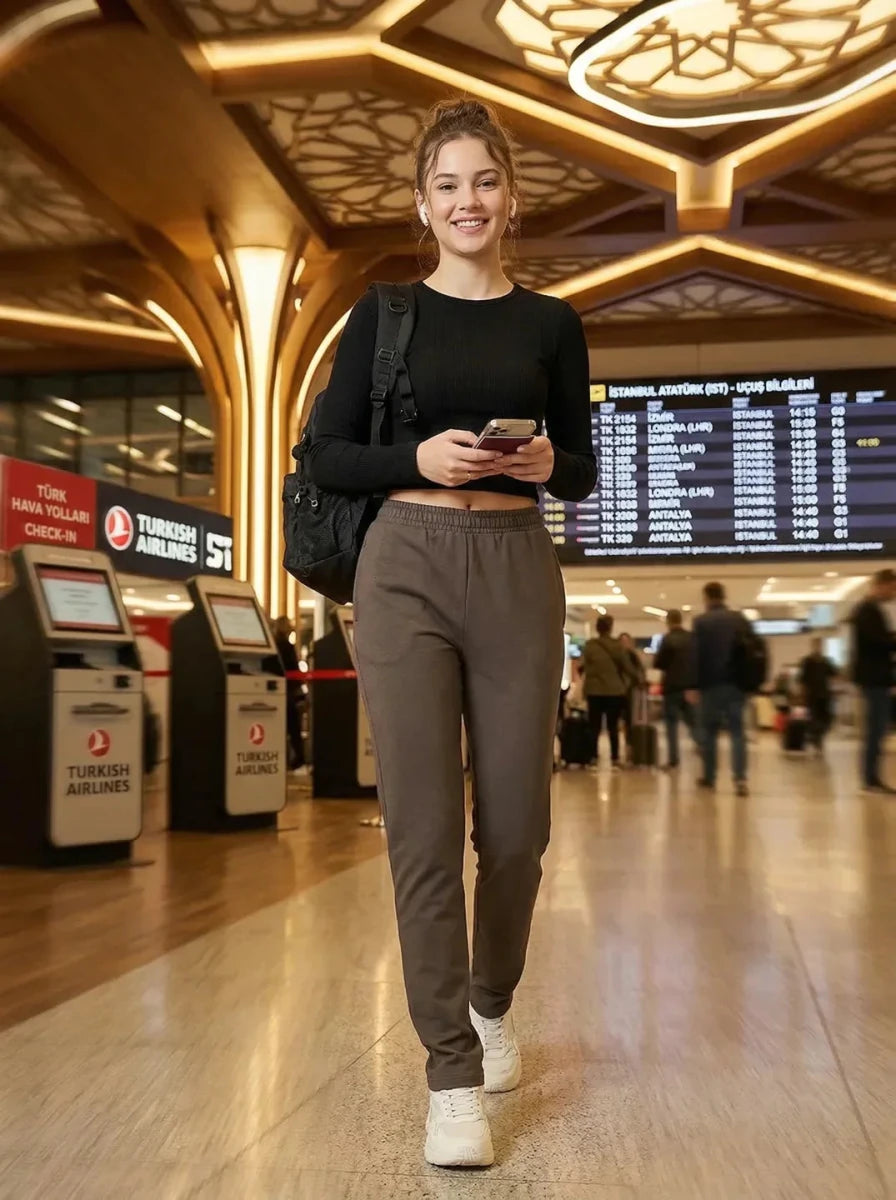 A woman in 98degreenorth's Olive Straight Fit Cotton Trackpants walks at the airport, phone in hand, flight boards behind.