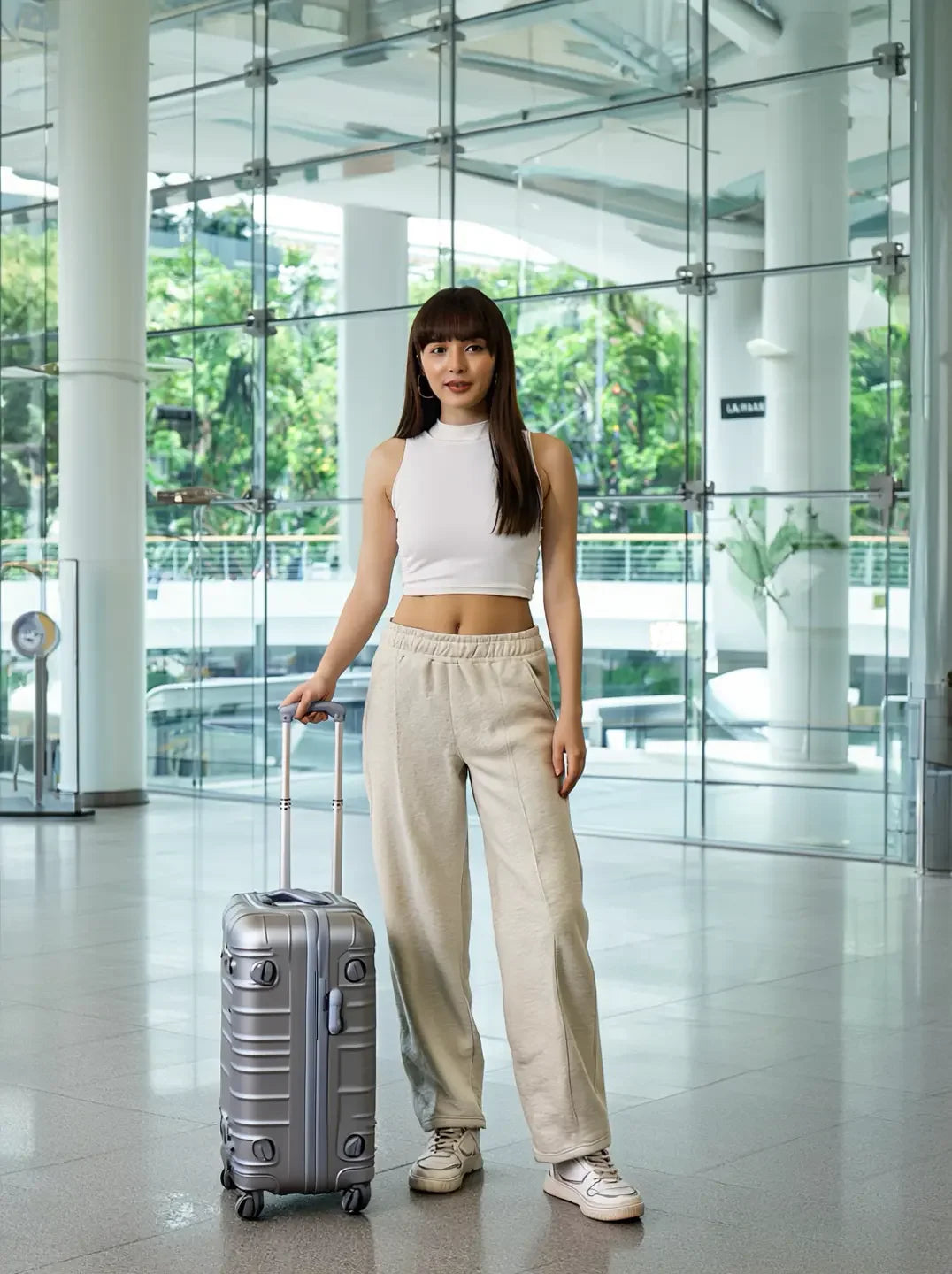 Woman in 98degreenorth Women’s Straight Fit Fleece Sweatpants in Ecru stands indoors with a silver suitcase, greenery outside.