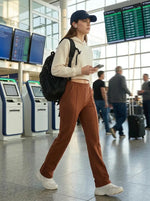 A woman in 98degreenorth Women's Tailored Straight Leg Pants in Rust walks through an airport, phone and backpack in hand.