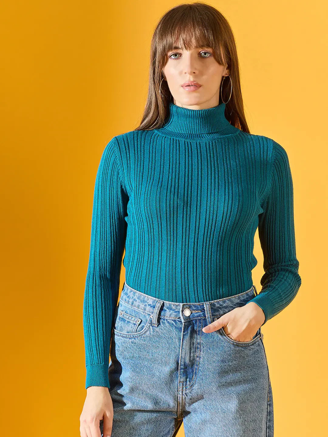 Woman wears 98degreenorth Teal Blue Ribbed Turtleneck Sweater, styled with jeans on a yellow backdrop. Winter fashion India.