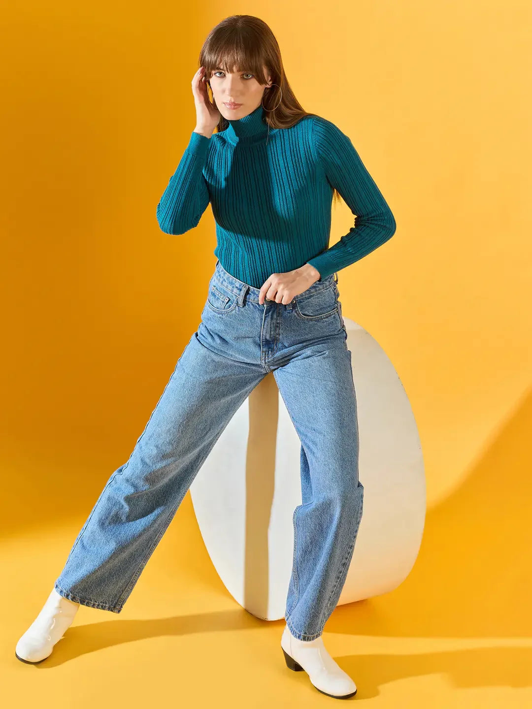 Woman models 98degreenorth’s teal blue ribbed turtleneck sweater against a bright yellow background.