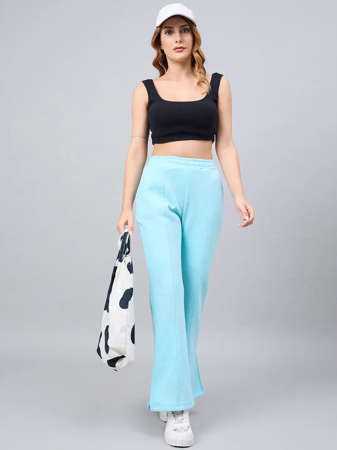 Woman in a black crop top, 98degreenorth turquoise flared bootcut sweatpants, white sneakers, cap, and a tote bag.