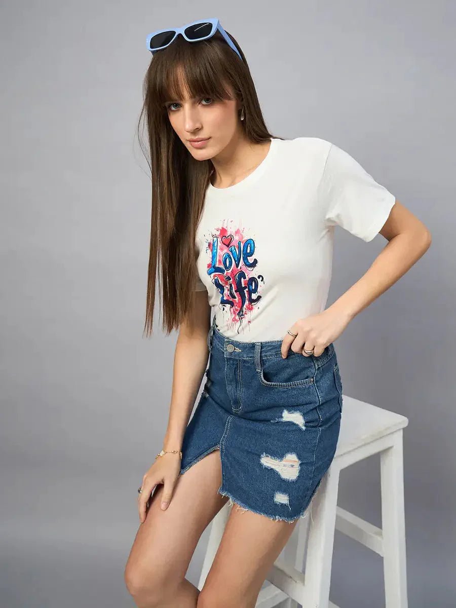 Woman in a 98degreenorth Women’s Typography Printed Cotton T-shirt and ripped denim skirt sits on a white stool.