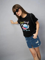 Woman in sunglasses wears the 98degreenorth Women’s Typography Printed Cotton Tee with a ripped denim skirt against a gray backdrop.