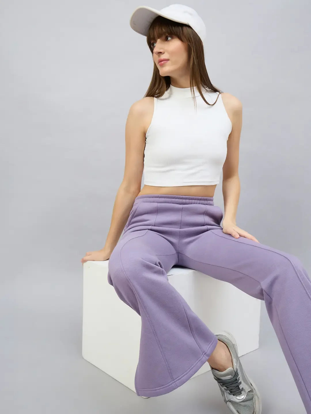 Woman in a white crop top and 98degreenorth Violet Flared Bootcut Sweatpants sits on a white cube against a gray background.