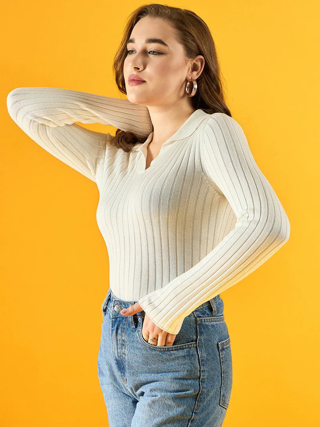 Woman wearing 98degreenorth Women’s White Ribbed Collar Neck Sweater and blue jeans poses by a yellow background.
