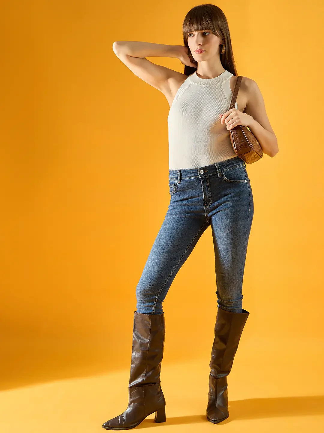 Woman wears 98degreenorth’s white ribbed halter neck sleeveless knit top, jeans, and brown boots, holding a small bag.