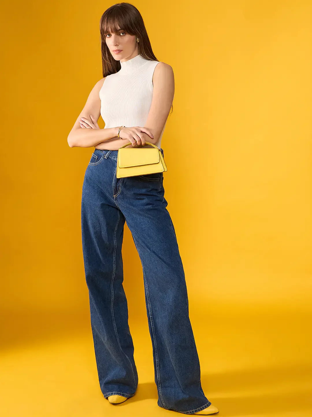 A woman in a 98degreenorth white ribbed high neck knit tank, wide-leg jeans, and yellow shoes holds a yellow handbag.