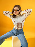Woman in 98degreenorth white ribbed striped turtleneck sweater and jeans poses against a bright yellow background.