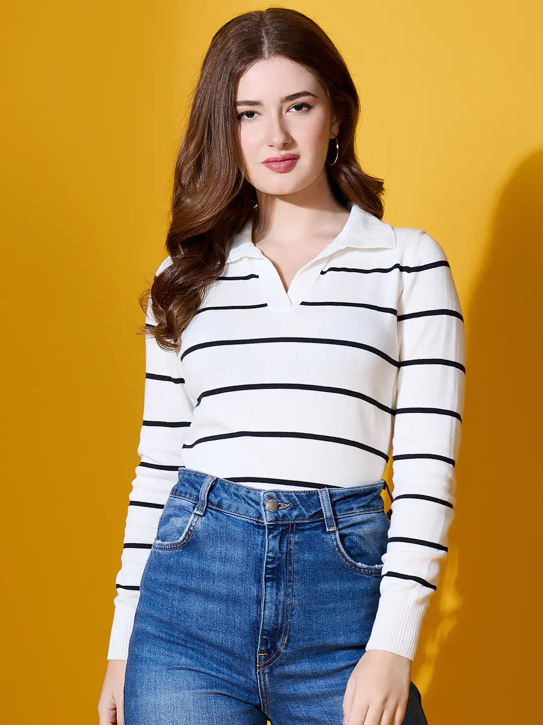 Woman in a 98degreenorth white striped collared knit sweater and blue jeans, standing against a yellow background.